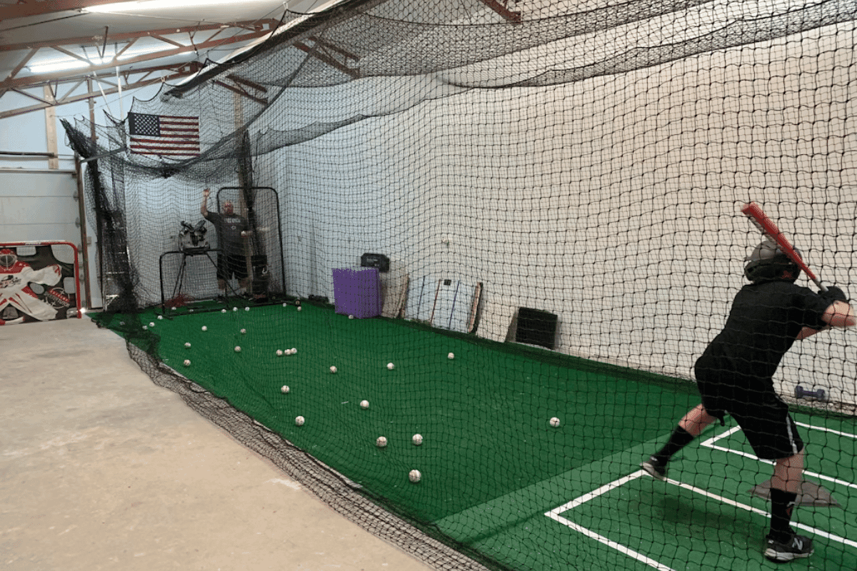A player takes a swing in an indoor batting cage as a coach feeds pitches