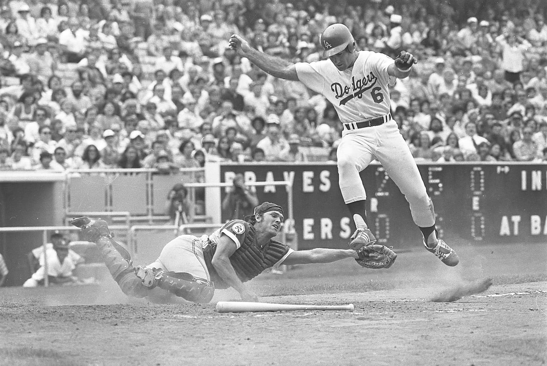 Black and white photo of Dodgers first baseman Steve Garvey leaping over the Atlanta catcher to score at home plate in 1976, with a packed crowd behind