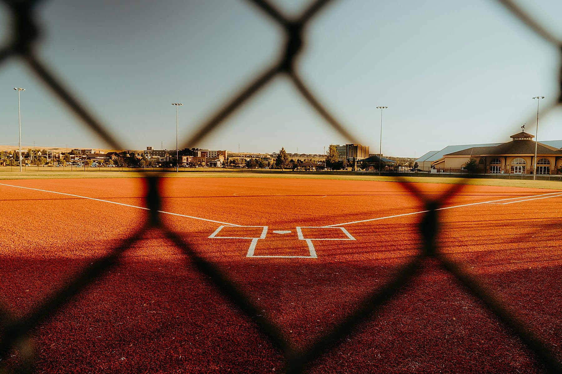 An empty baseball diamond at golden hour seen through a chain-link fence, with nobody behind home plate