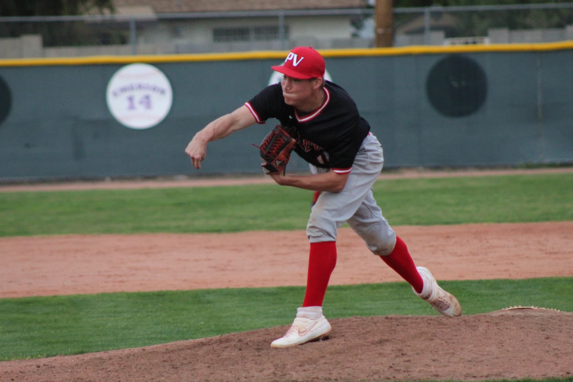 A high school pitcher in a red cap and black jersey follows through on a pitch from the mound during a game