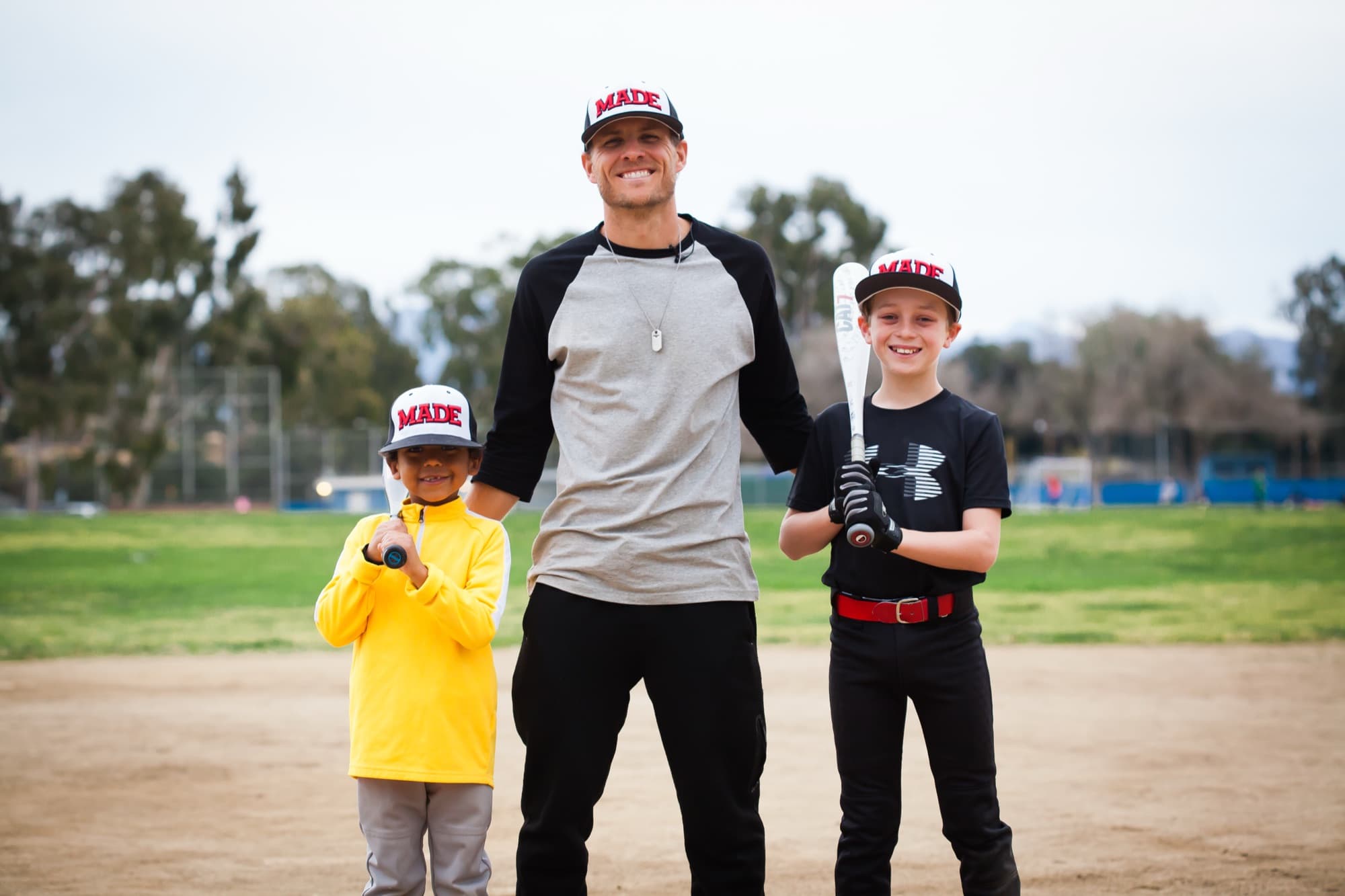 A coach standing with two young players holding bats on a youth baseball field