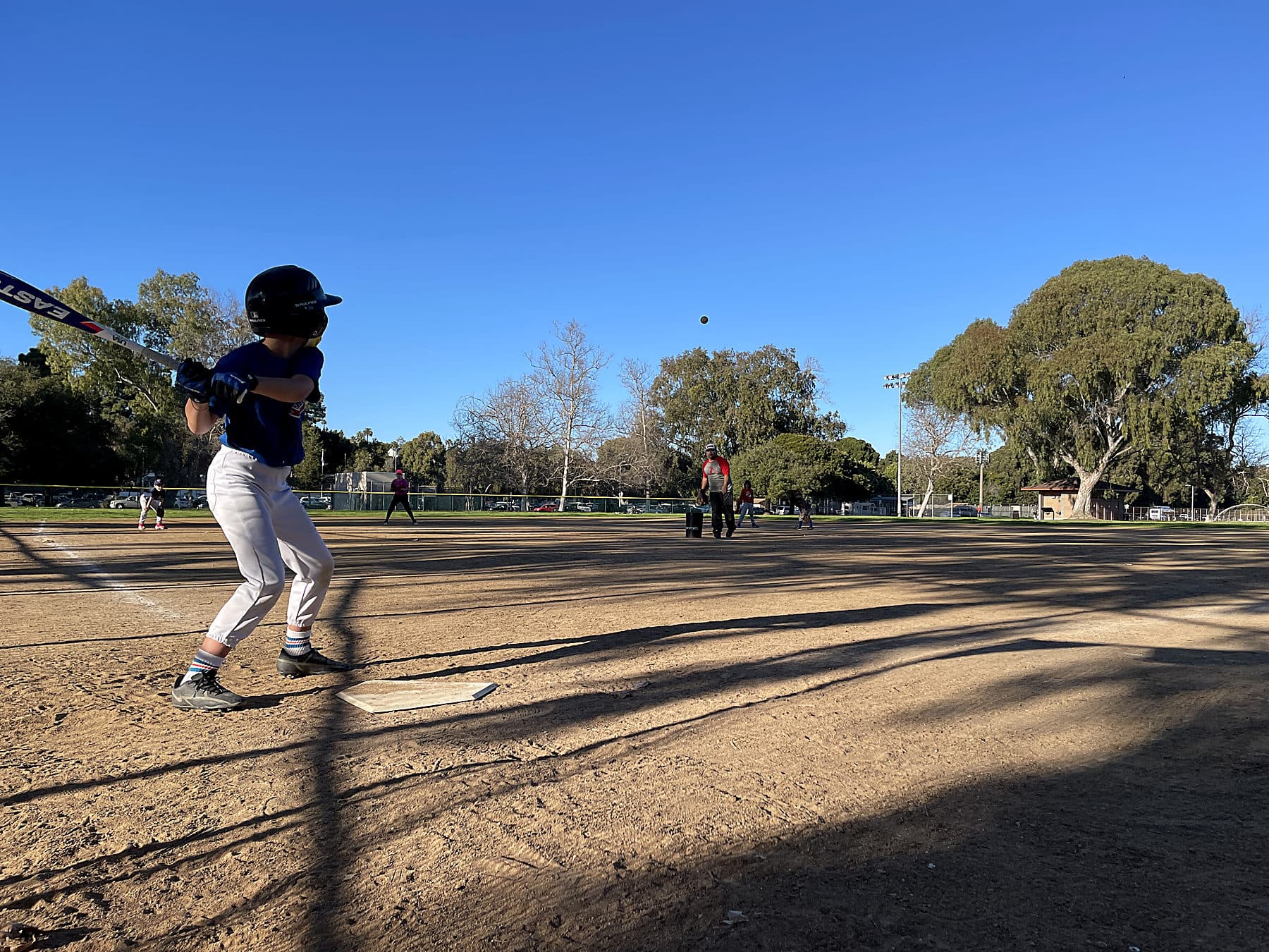 A young hitter in a batting stance at home plate at Penmar Park on a sunny afternoon, with the pitch in the air and a coach pitching from the mound