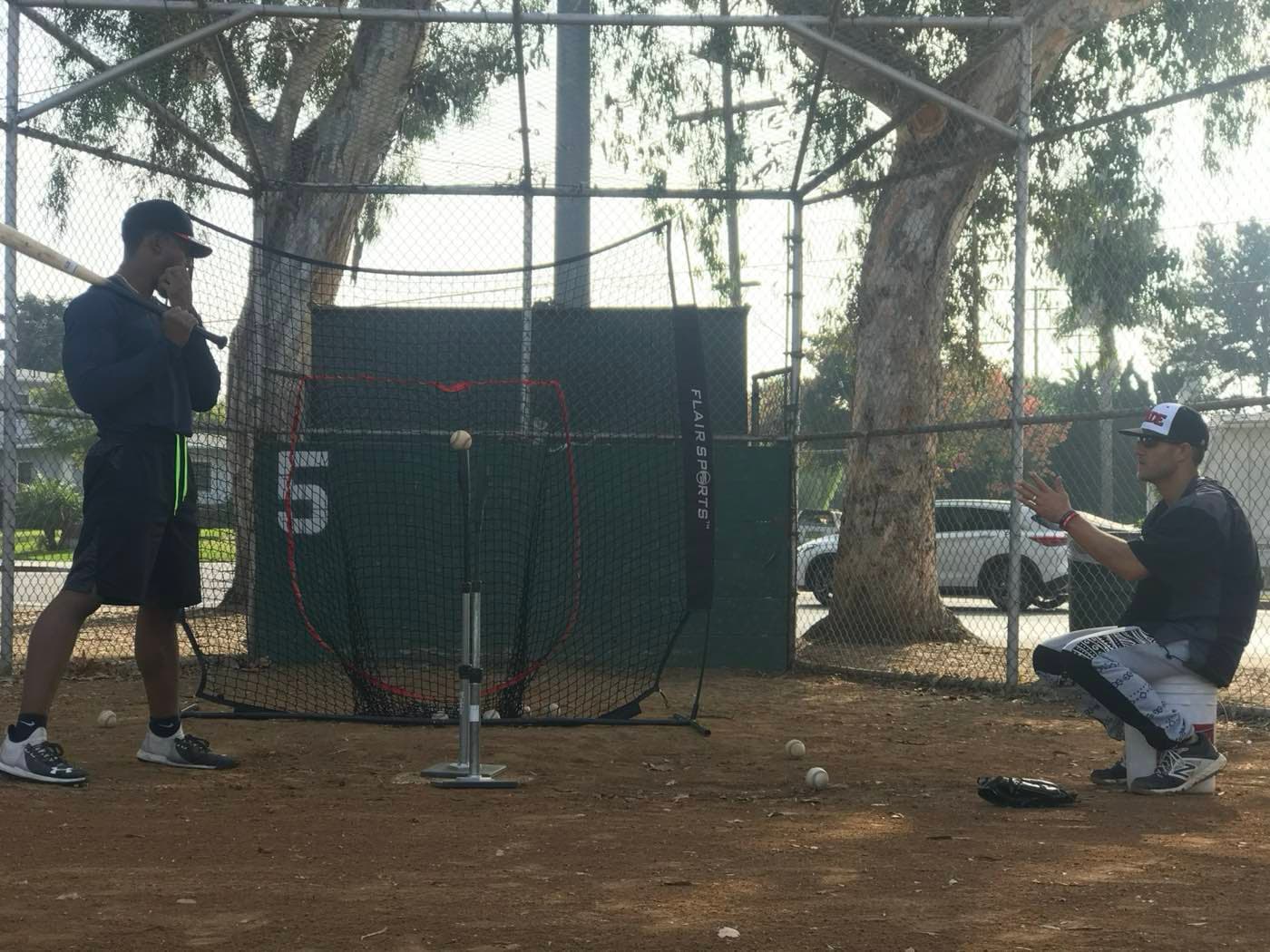 A hitter working off a tee into a net inside a fenced outdoor batting cage on a dirt ball field, with a coach front-tossing from one knee
