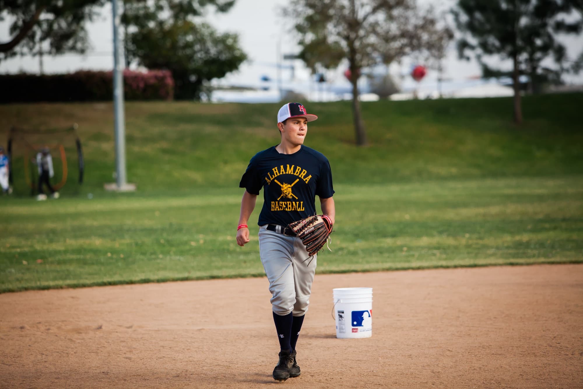 A youth baseball player in a glove walks across the infield dirt of a park field in the evening