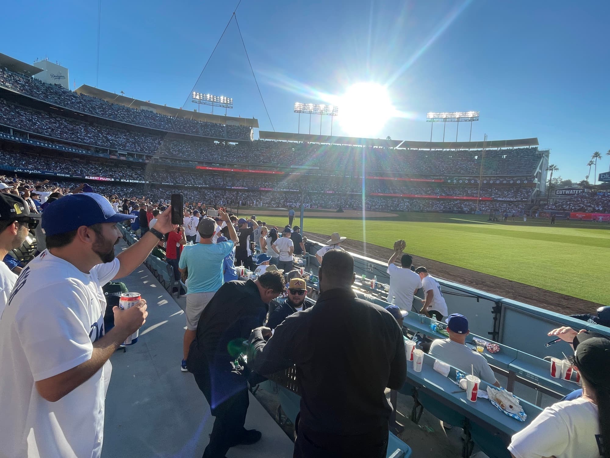 A ballpark seen from the outfield pavilion on a sunny afternoon, with the field, the stands and a bright sun flare