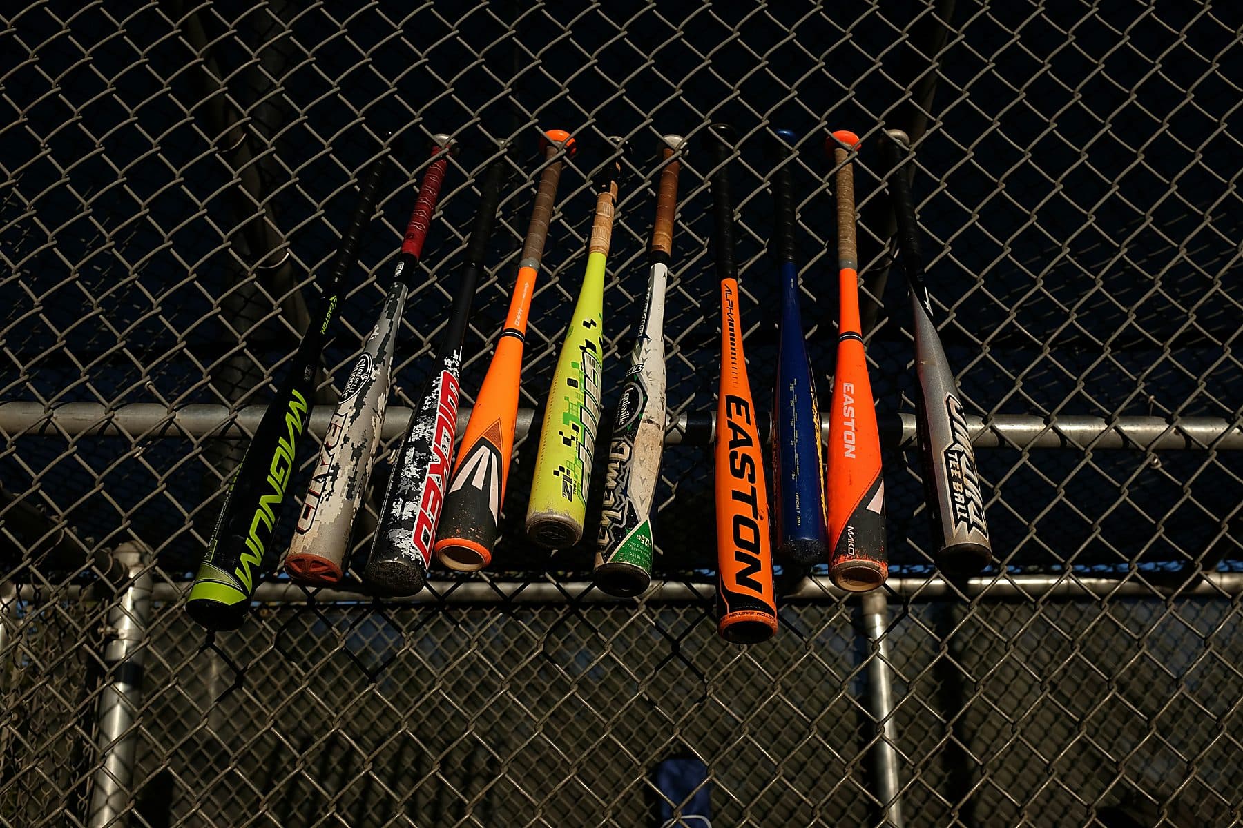 A row of premium composite baseball bats hanging on a dark chain-link fence