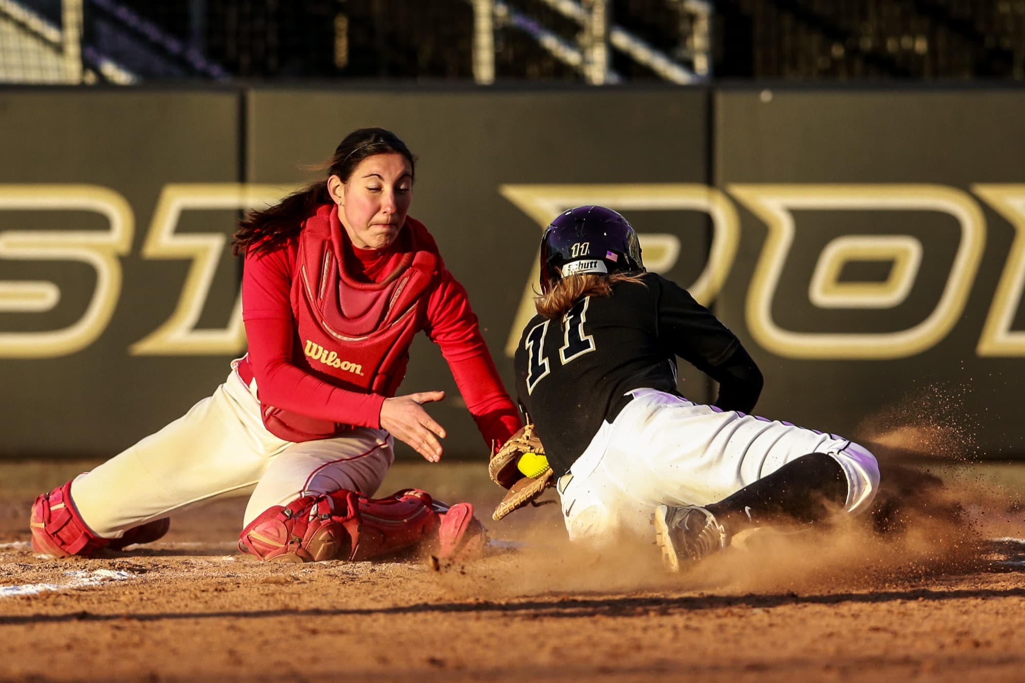 A college softball catcher in a red uniform reaches down with her glove to tag a sliding runner in a black uniform at home plate, with a yellow softball and dust in the air