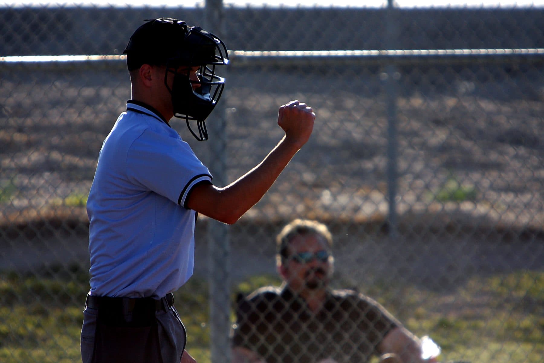A young volunteer umpire in mask and light blue shirt signals a strike with his fist while a spectator watches through the backstop fence
