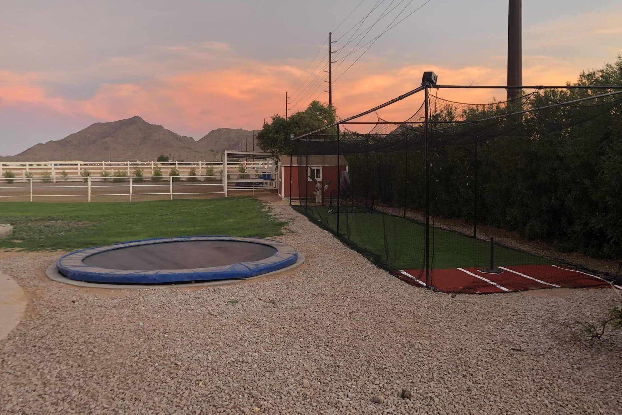 A backyard batting cage at sunset next to a sunken trampoline, desert mountains in the distance