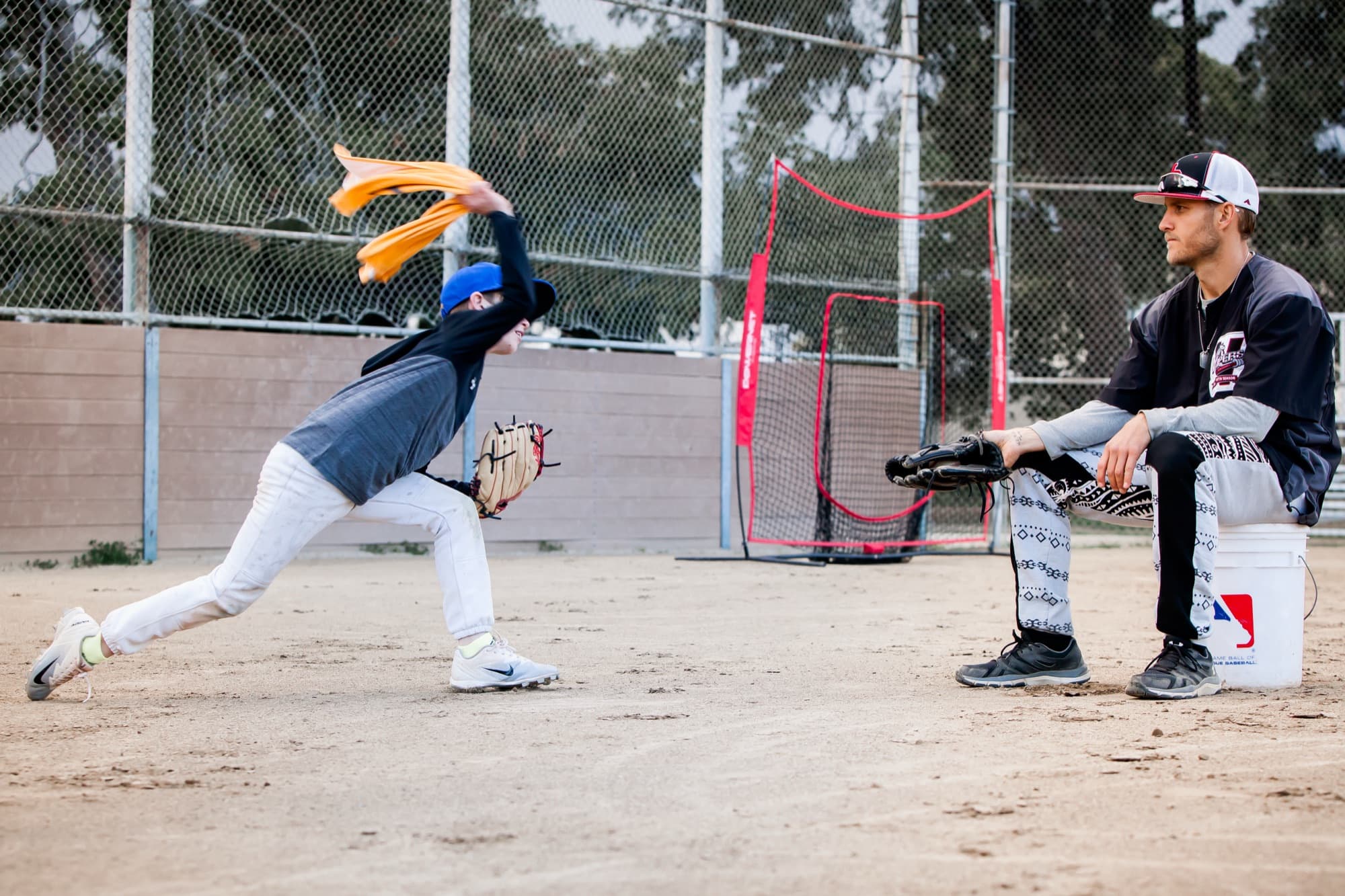 A young pitcher swings a towel through his throwing motion on a dirt field while a coach sits on a bucket with a glove, watching