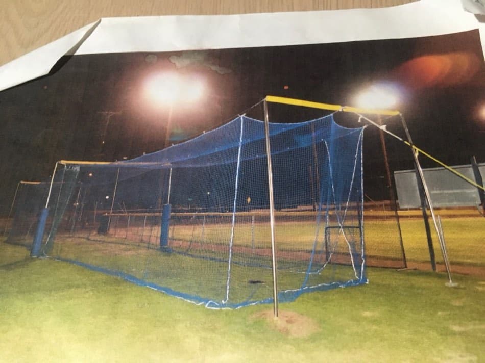 A blue-netting batting cage lit up at night on a ballfield