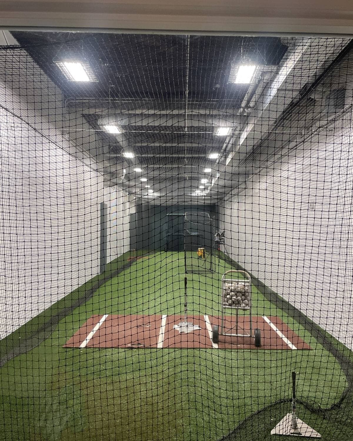 An indoor batting tunnel with turf, a home plate mat, a tee and a cart of baseballs