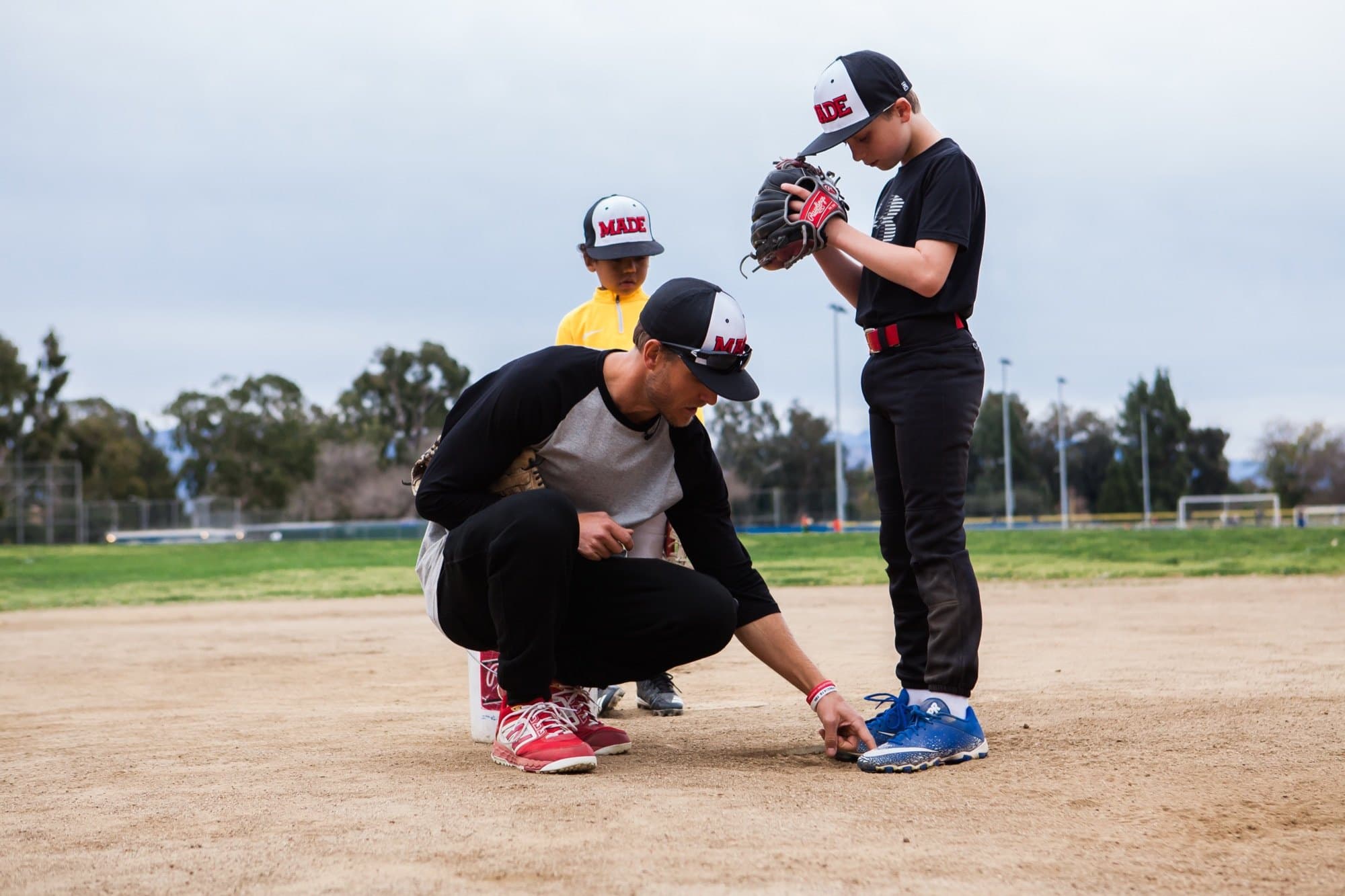 A coach crouched beside a young player on an infield, setting the player's feet