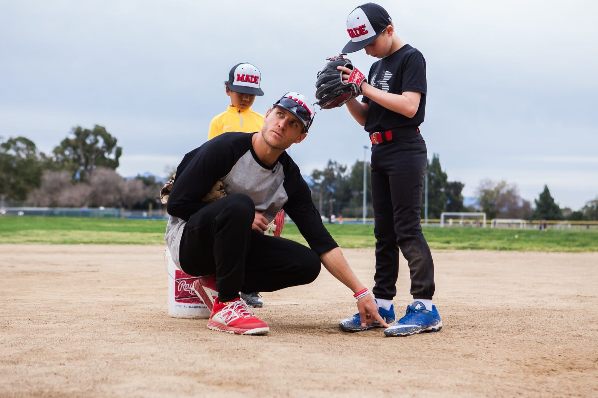 A coach kneeling on an infield, pointing at a young player's front foot while a teammate watches