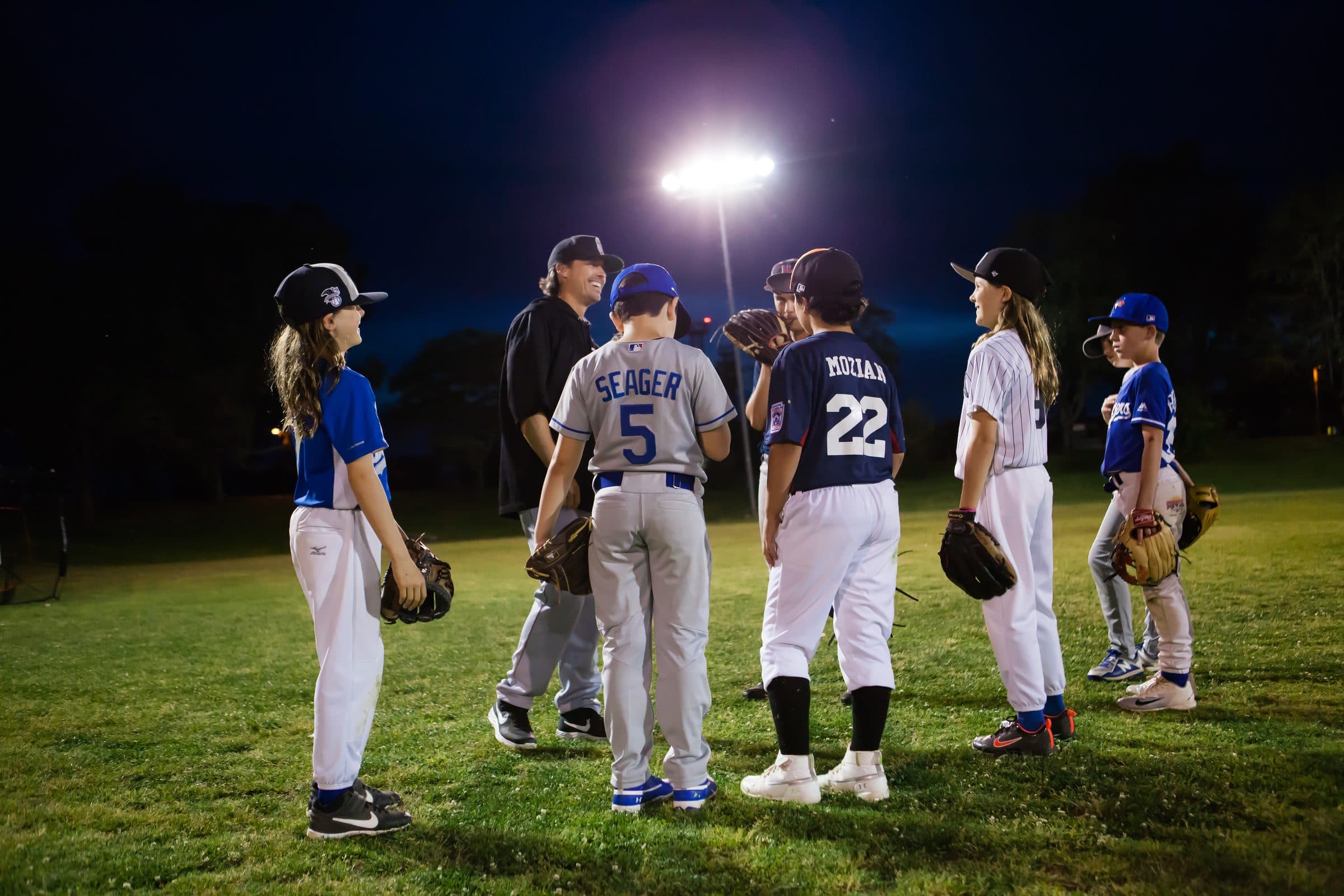 A coach under the field lights talking with a group of young players holding their gloves