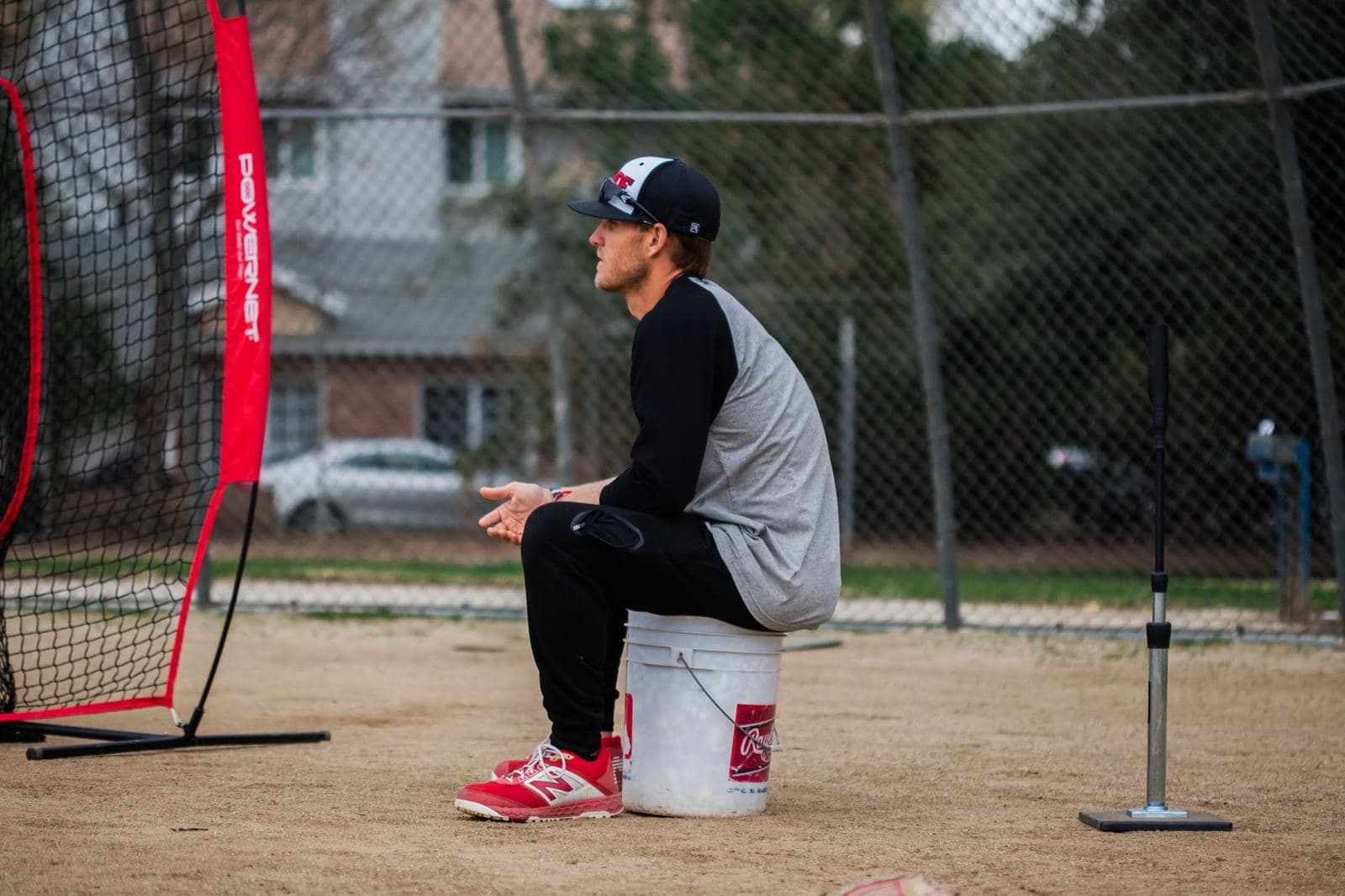 Coach Isaac Hess sitting on a ball bucket beside a batting tee and hitting net