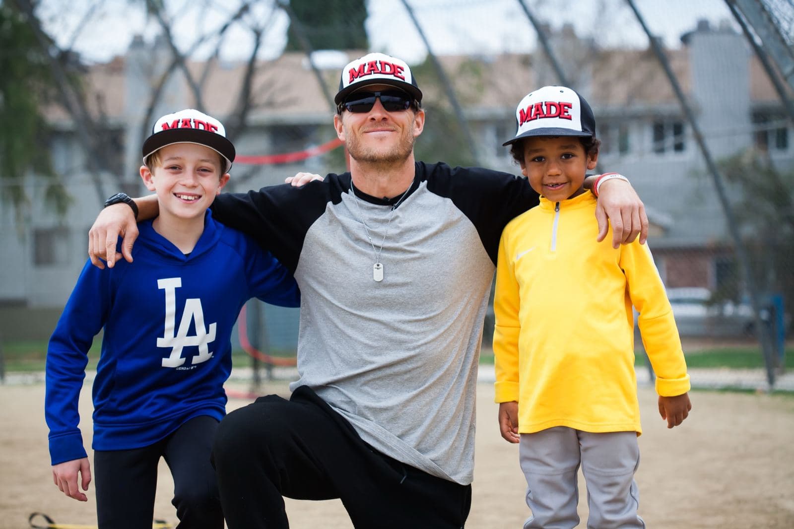 Coach Isaac Hess kneeling with an arm around two smiling young players in MADE Baseball caps