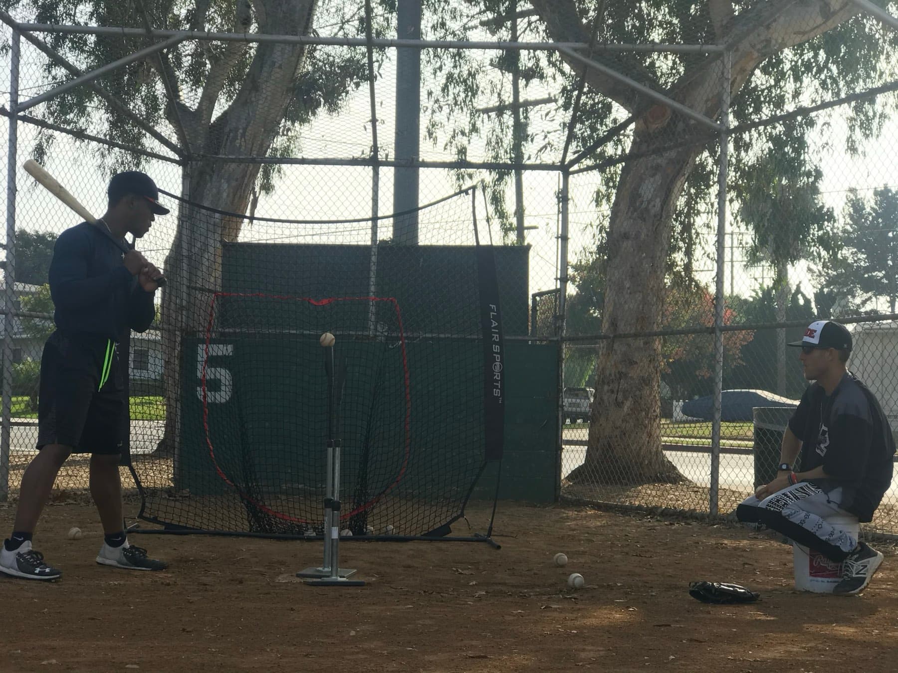 A hitter loading up at the tee in an outdoor cage while a coach watches from a bucket