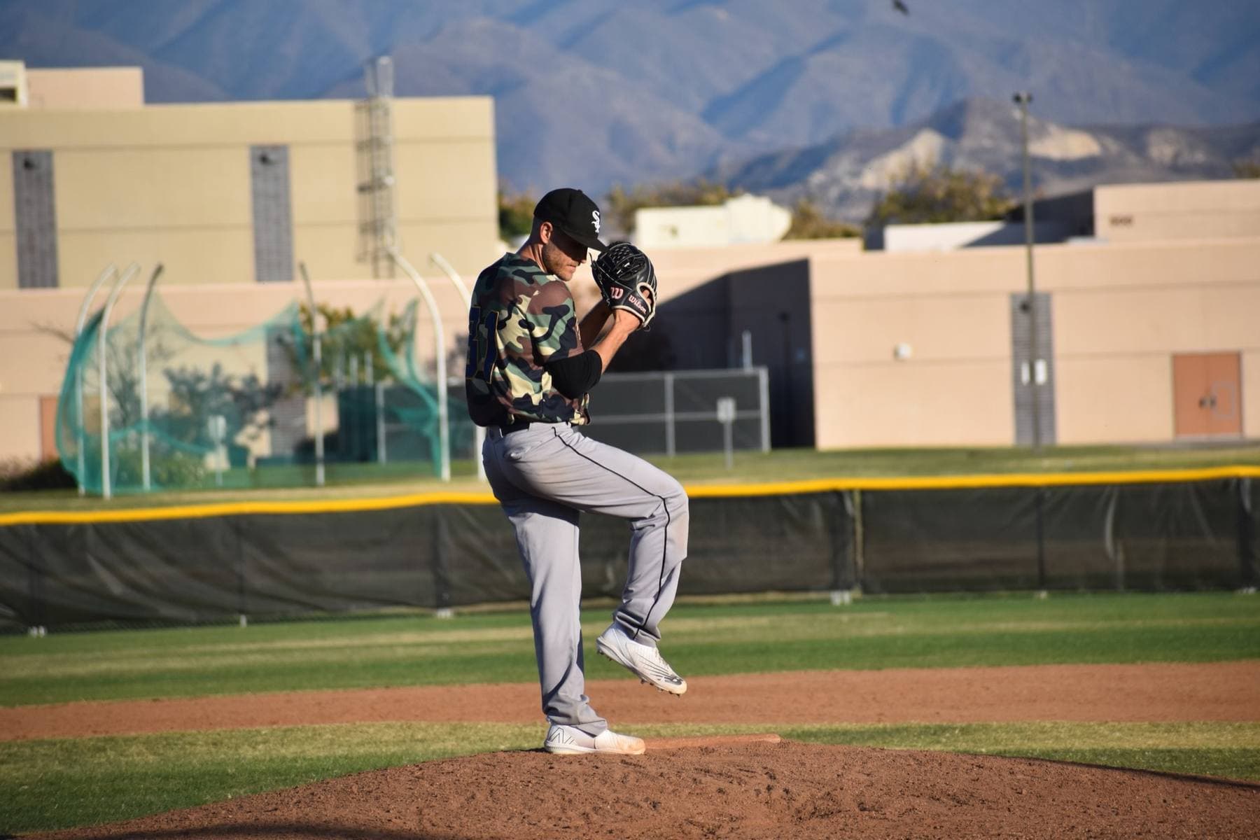 An adult league pitcher at the top of his delivery, mountains behind the outfield fence