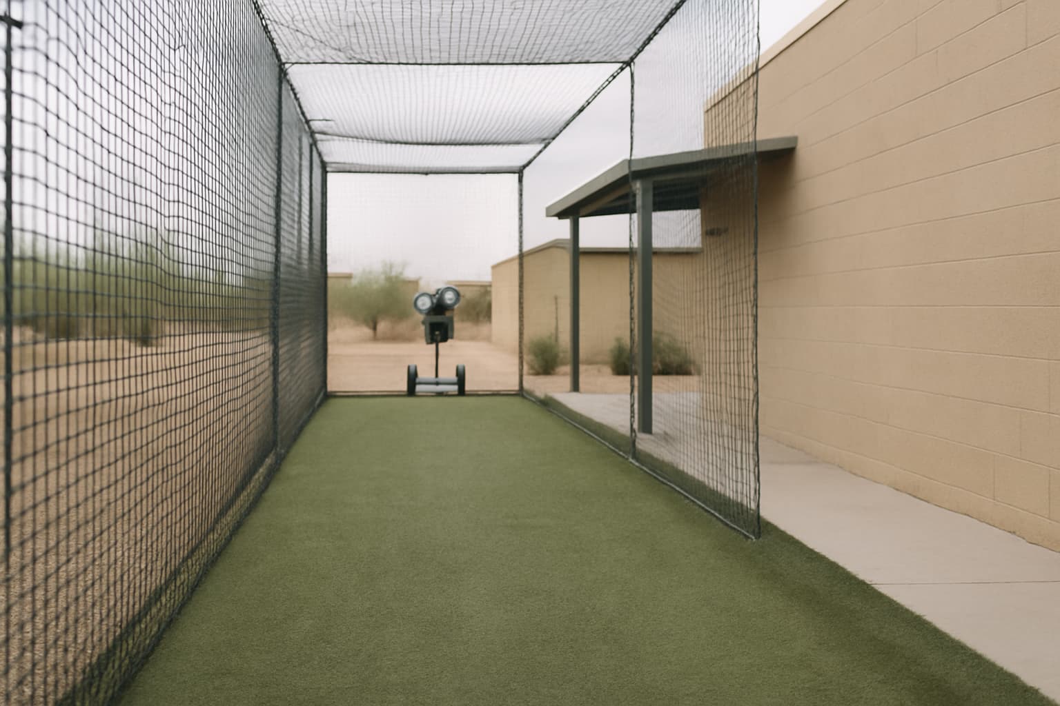 Empty batting cage with a pitching machine in the background and dry landscaping.