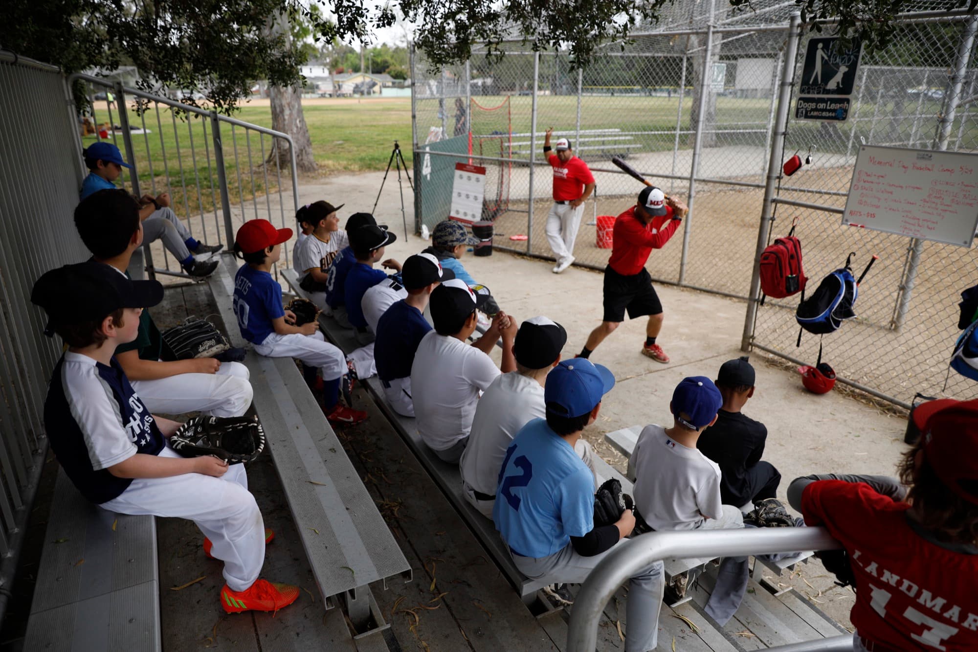 Young players in mixed club jerseys sit on a dugout bench watching a MADE Baseball coach demonstrate a swing at a paid camp.