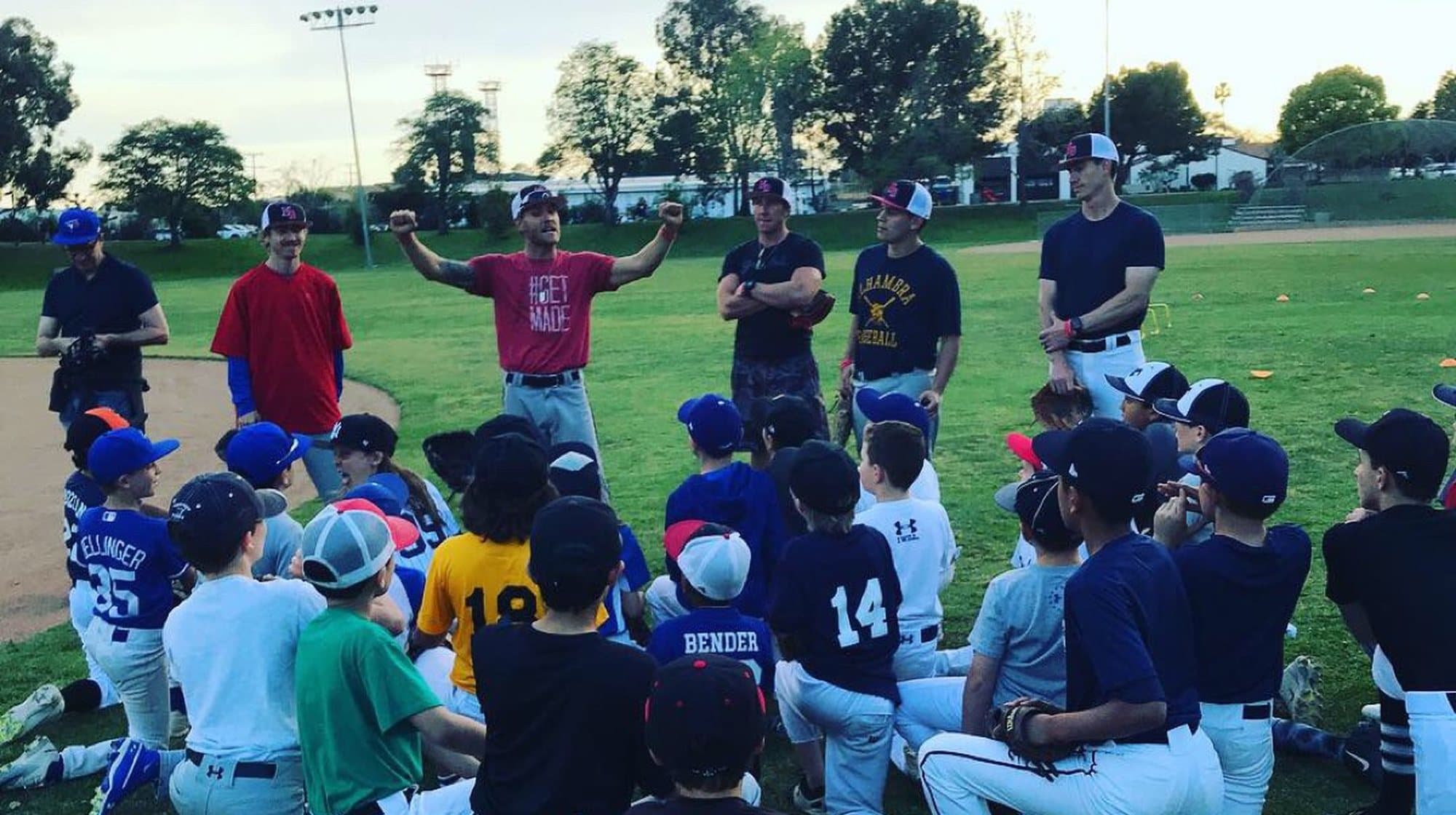 Isaac Hess in a MADE Baseball shirt with his arms raised, addressing a semicircle of kneeling young players and five other coaches at dusk.