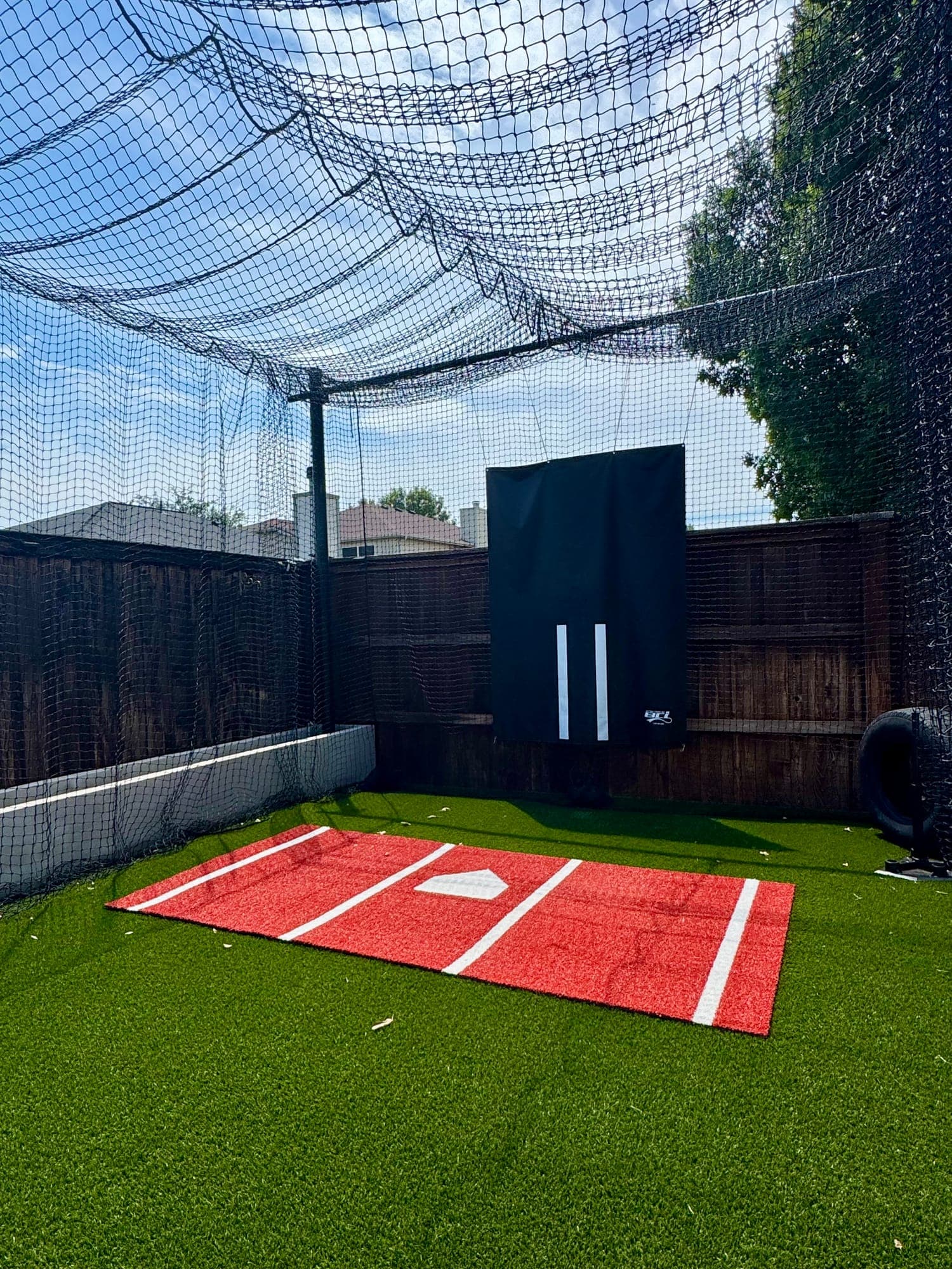 Bright backyard cage with red hitting mat, fresh green turf and black backdrop