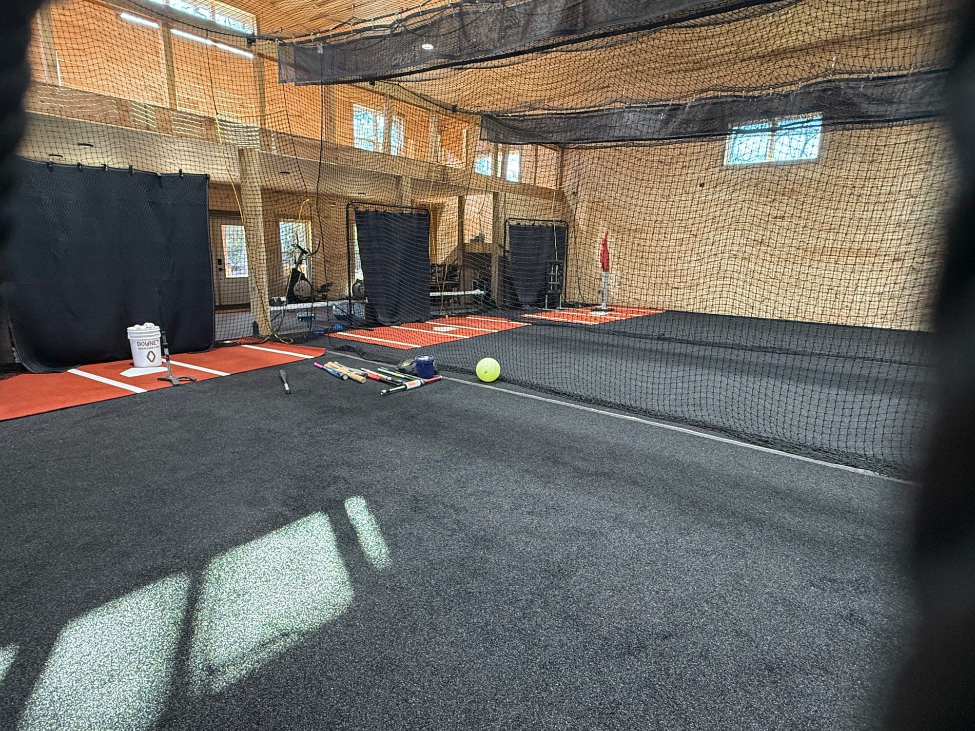 Netted indoor cage with dark carpeted floor, red mats, bats and a yellow softball