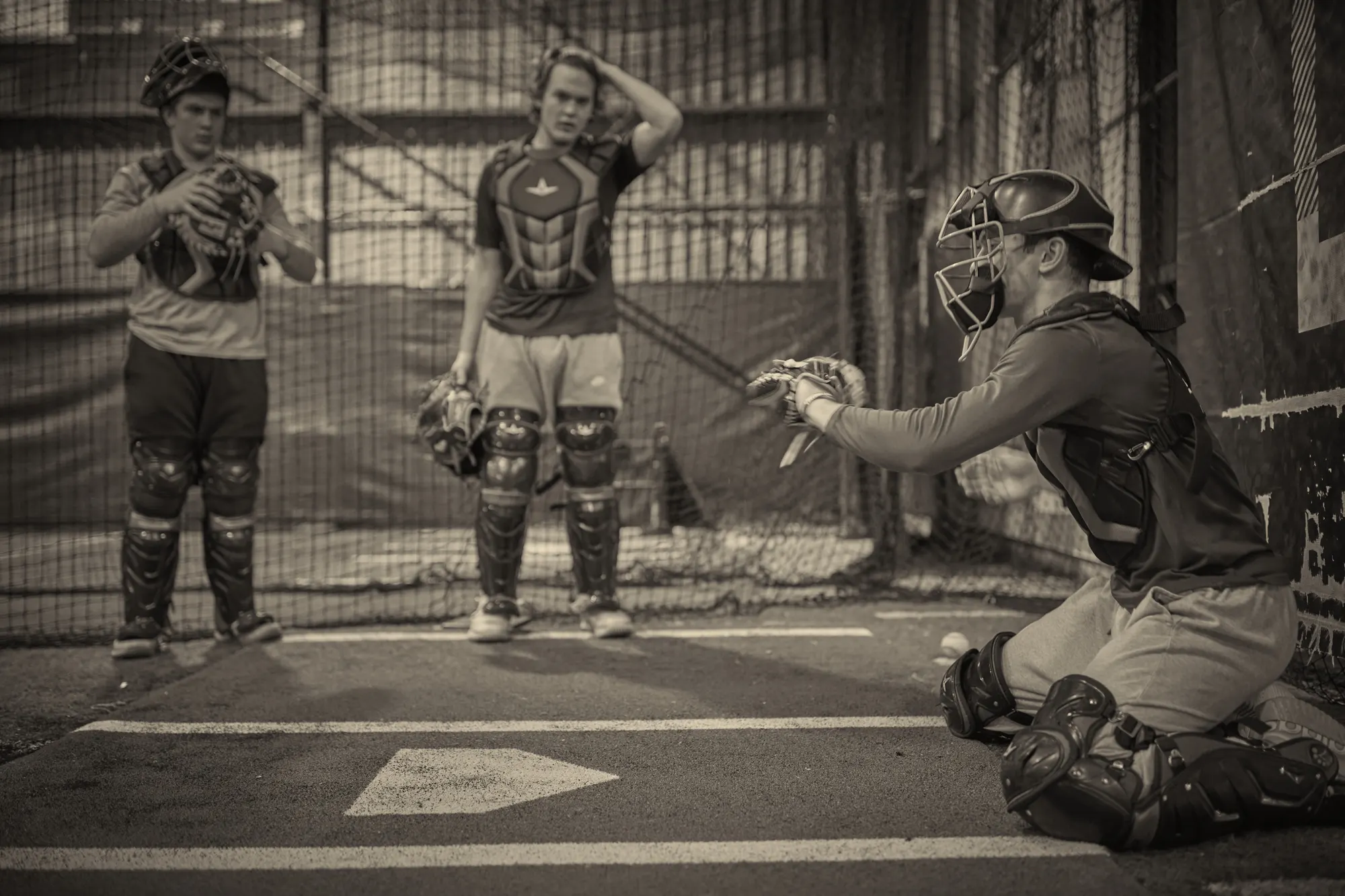 Three catchers in full gear working at home plate inside a batting cage