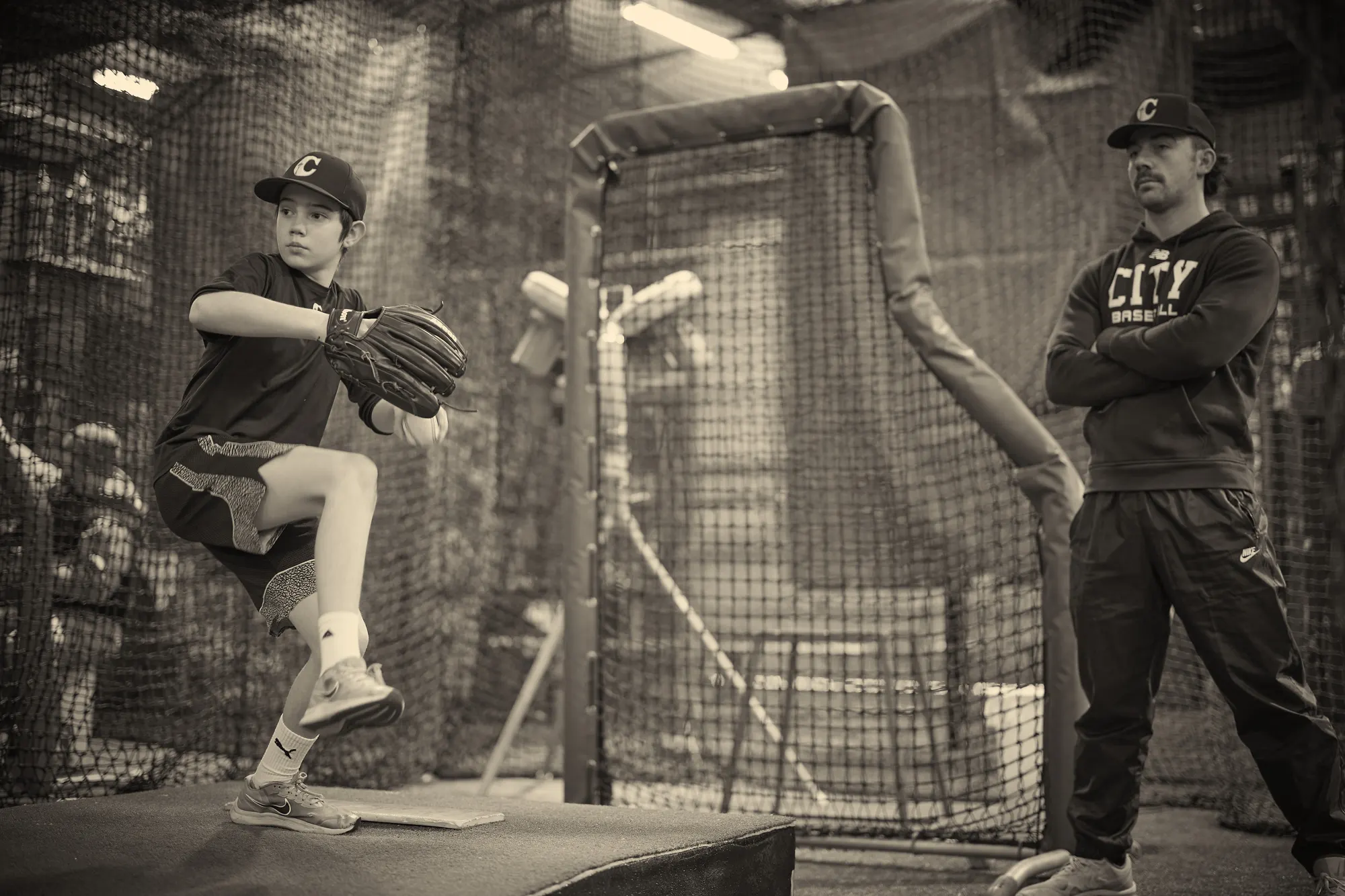A young pitcher mid-windup on an indoor mound while a coach in a City Baseball hoodie watches
